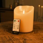 Ładowalna świeca LED z wosku Ø12,5 × 15 cm – ciepłe białe – na drewnianym stole z pilotem DORSETTO, w tle rozmyte świeczki