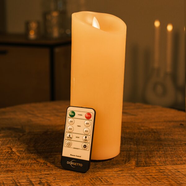 Ładowalna świeca LED z wosku DORSETTO Ø7,5 × 20 cm na drewnianym stole z pilotem, przytulne oświetlenie i rozmyte świece w tle