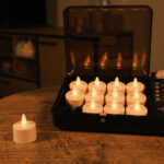 12 ładowalnych, wodoodpornych podgrzewaczy LED DORSETTO w czarnym etui ładującym na drewnianym stole – jeden podgrzewacz poza etui, w tle delikatnie oświetlony pokój