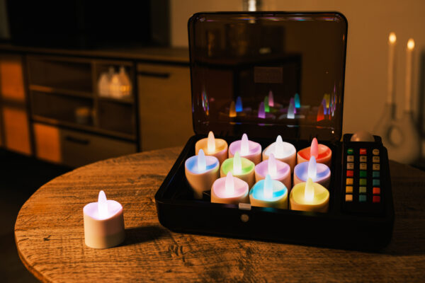 Zestaw 12 podgrzewaczy LED RGB DORSETTO w otwartym czarnym etui na okrągłym drewnianym stole – jeden podgrzewacz ustawiony poza etui, w tle nowoczesny salon