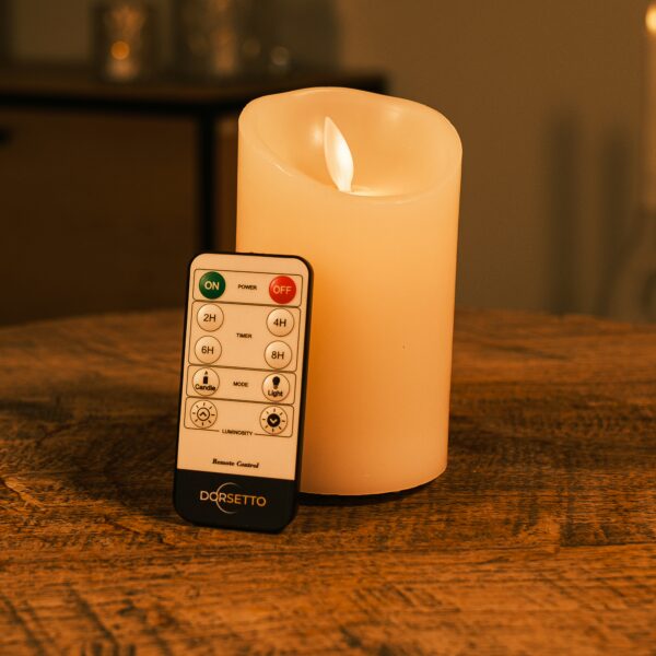 Ładowalna świeca LED z wosku DORSETTO z ruchomym płomieniem Ø 7,5 × 12,5 cm – na drewnianej powierzchni obok pilota z funkcją timera i regulacją jasności