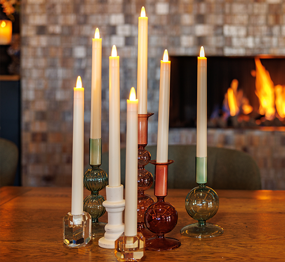 Een groep van acht brandende kaarsen in kleurrijke glazen en kristallen houders staat op een houten tafel, met een open haard en warm, gezellig interieur op de achtergrond.