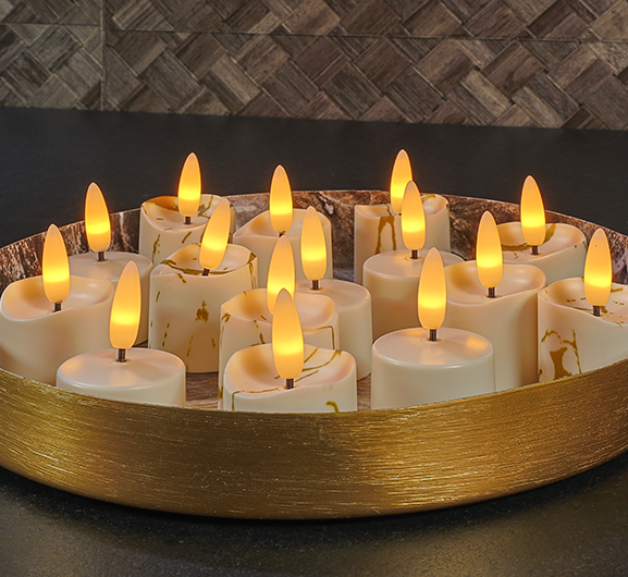 Een rond gouden dienblad bevat verschillende brandende, vlamloze LED-kaarsen met wit en goud gemarmerde buitenkant, dicht bij elkaar geplaatst op een donker oppervlak tegen een gestructureerde achtergrond.