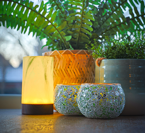 Een zacht gloeiende kaarslamp staat op een tafel naast twee theelichthouders van mozaïekglas en groene planten in potten en creëert een gezellige, warme sfeer bij een raam.