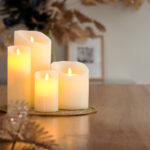 Zestaw świec LED z wosku DORSETTO (4 szt.) świecących ciepłym światłem na drewnianym stole, ustawionych na złotej tacy z dekoracyjnymi liśćmi – przytulna i świąteczna atmosfera