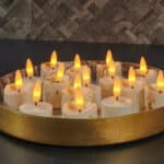 Okrągła złota taca z kilkoma tealightami LED z płomieniem 3D, ładowalnymi, z pilotem i funkcją timera, ustawiona na ciemnym blacie; w tle teksturowana ściana z płytek.