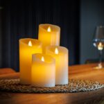 Zestaw 4 ładowalnych świec LED z wosku DORSETTO o różnych wysokościach na plecionej macie na drewnianym stole – ciepłe światło z kieliszkiem wina i butelką w tle
