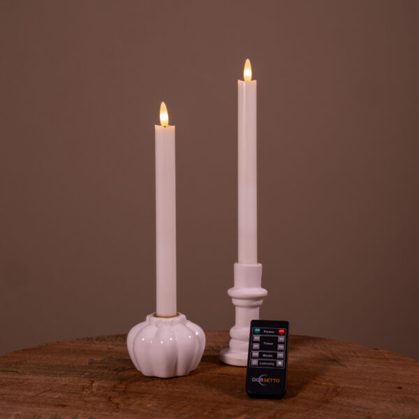 Zestaw 2 białych świec stołowych LED (400 godzin, ładowalne) w ceramicznych świecznikach – jeden okrągły z fakturą, drugi klasyczny – ustawione na drewnie, z pilotem z przodu.