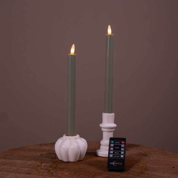 Dwie zapalone świece stołowe LED (400 godzin świecenia, ładowalne, zestaw 2 szt.) stoją w białych ceramicznych świecznikach na drewnianej powierzchni, a z przodu leży mały czarny pilot Dorsetto. Tło jest jednolicie brązowe.