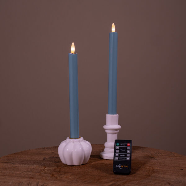 Dwie świece stołowe LED w kolorze kamiennego niebieskiego (zestaw 2, do 400 godzin świecenia) w białych świecznikach na drewnianej powierzchni, z czarnym pilotem z przodu i jednolitym brązowym tłem.
