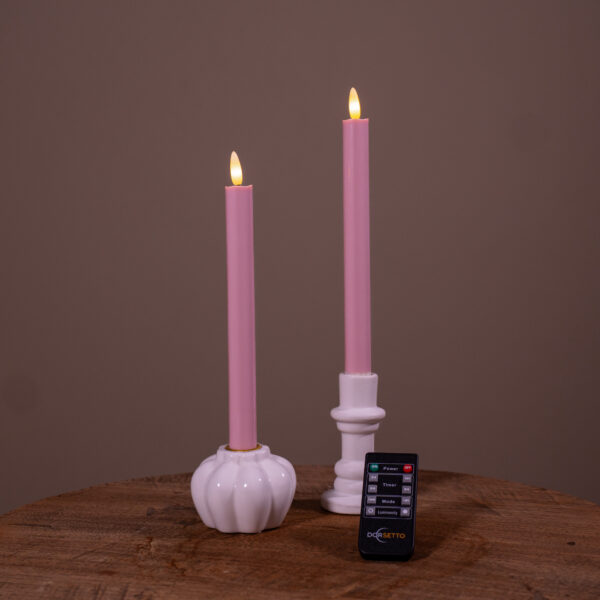 Dwie świece stołowe LED (400 godzin, ładowalne) w kolorze pudrowego różu – zestaw 2 szt. – stoją w białych świecznikach na drewnianej powierzchni. Z przodu leży mały pilot. Tło jest jednolicie brązowe. html Copy code