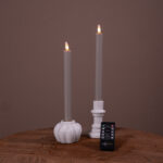 Dwie ciemnoszare świece stołowe LED (ładowalne, do 400 godzin świecenia, zestaw 2 szt.) w białych świecznikach na drewnianej powierzchni, z małym pilotem DORSETTO z przodu i jednolitym brązowym tłem.