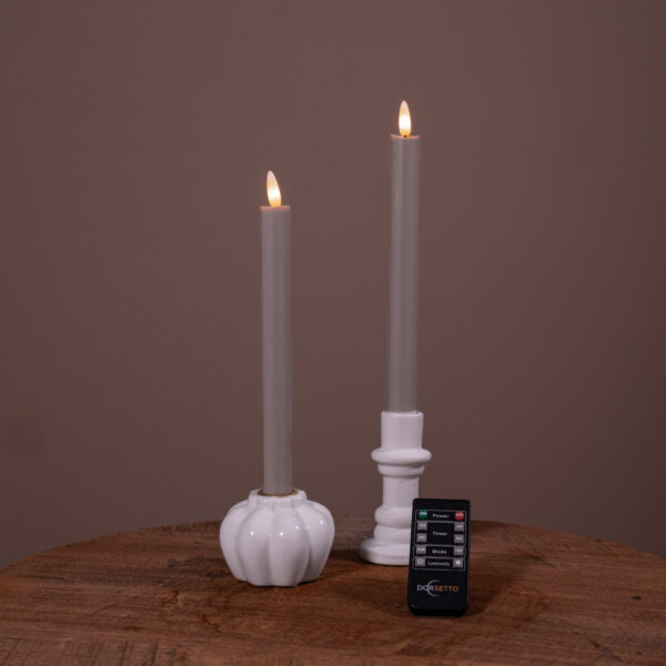 Dwie ciemnoszare świece stołowe LED (ładowalne, do 400 godzin świecenia, zestaw 2 szt.) w białych świecznikach na drewnianej powierzchni, z małym pilotem DORSETTO z przodu i jednolitym brązowym tłem.