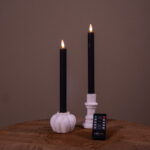 Dwie beżowe ładowalne świece stołowe LED (zestaw 2 szt., do 400 godzin świecenia) umieszczone w białych świecznikach – jednym w kształcie dyni, drugim cylindrycznym – stoją na drewnianej powierzchni, z pilotem z przodu, na jednolitym tle.