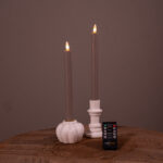 Dwie beżowe świece stołowe LED (zestaw 2 szt., do 400 godzin świecenia, ładowalne) stoją w białych świecznikach na drewnianej powierzchni, z małym czarnym pilotem z przodu. Jednolite brązowe tło dopełnia całość.