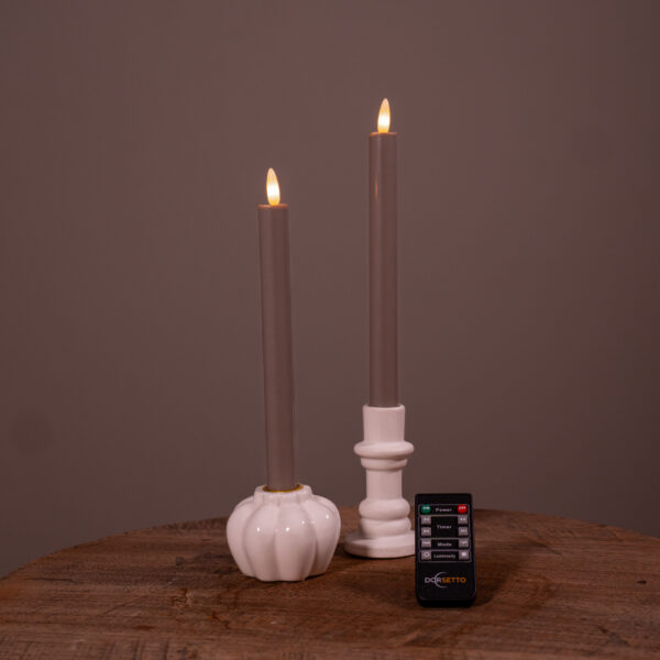Dwie beżowe świece stołowe LED (zestaw 2 szt., do 400 godzin świecenia, ładowalne) stoją w białych świecznikach na drewnianej powierzchni, z małym czarnym pilotem z przodu. Jednolite brązowe tło dopełnia całość.