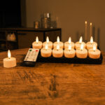 Dwanaście ładowalnych tealightów z realistycznymi płomieniami 3D, do 140 godzin świecenia i pilotem stoi na drewnianym stole, tworząc delikatną, przytulną poświatę w tle.