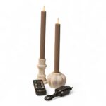 Dwie brązowe, bezpłomieniowe stożkowe świece LED w białych świecznikach, pokazane wraz z czarnym pilotem i czarnym zasilaczem, na jednolitym białym tle.