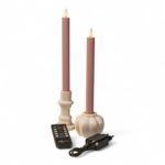 Dwie różowe świece LED na baterie w białych świecznikach, pilot oraz czarny zasilacz na białym tle. Świece mają końcówki w kształcie płomienia imitujące prawdziwy ogień.