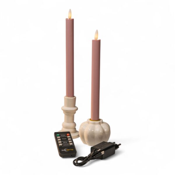 Dwie różowe świece LED na baterie w białych świecznikach, pilot oraz czarny zasilacz na białym tle. Świece mają końcówki w kształcie płomienia imitujące prawdziwy ogień.