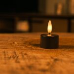 Dwanaście czarnych, ładowalnych tealightów o czasie świecenia do 400 godzin z pilotem stoi na drewnianej powierzchni i emituje ciepły blask. Delikatnie rozmyte tło tworzy przytulną, relaksującą atmosferę.