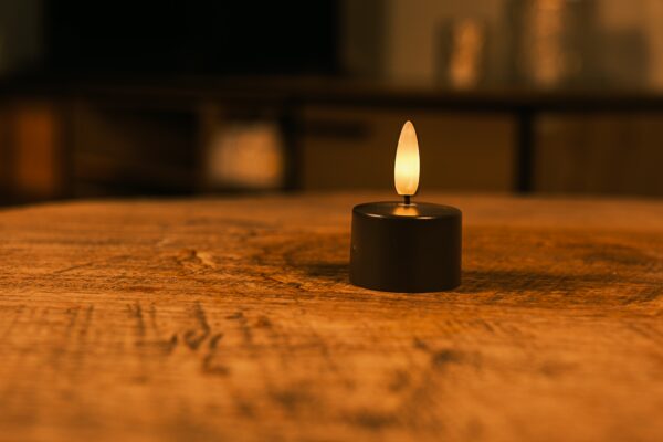 Dwanaście czarnych, ładowalnych tealightów o czasie świecenia do 400 godzin z pilotem stoi na drewnianej powierzchni i emituje ciepły blask. Delikatnie rozmyte tło tworzy przytulną, relaksującą atmosferę.