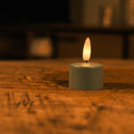 Zestaw 12 oliwkowych LED tealightów z pilotem stoi na drewnianej powierzchni, emituje ciepły, delikatny blask do 400 godzin i tworzy przytulną, intymną atmosferę.