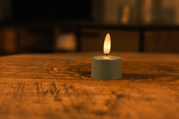 Zestaw 12 oliwkowych LED tealightów z pilotem stoi na drewnianej powierzchni, emituje ciepły, delikatny blask do 400 godzin i tworzy przytulną, intymną atmosferę.