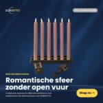 Zestaw sześciu różowych, ładowanych, ściemnianych świec obiadowych LED o średnicy 250 mm z pilotem, prezentowany przez Dorsetto. Holenderski tekst podkreśla romantyczną atmosferę bez otwartego ognia. Przycisk "Shop nu" pojawia się w prawym dolnym rogu. Produkt: Różowe świece obiadowe LED 250 mm - zestaw 6 szt, ładowalne, ściemnialne, z pilotem.
